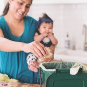 Woman holding baby adding food scraps to a green container.