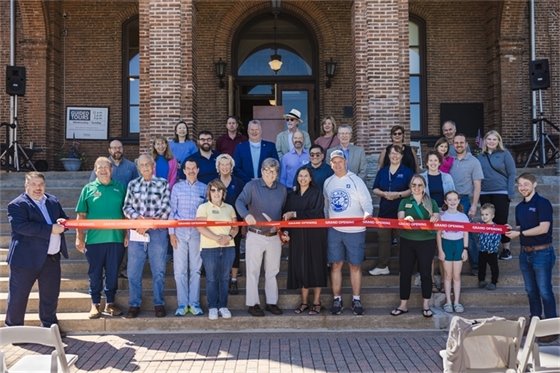 Group of people gathered for a ribbon-cutting ceremony in front of a brick building, with several individuals holding a large red ribbon.