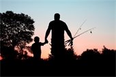 Silhouette of two individuals, one adult and one child, holding fishing rods against a vivid sunset sky with hues of orange and blue. The background shows a clear, tree-lined horizon.