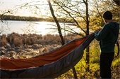 A man setting up a hammock on a tree in front of a lake.